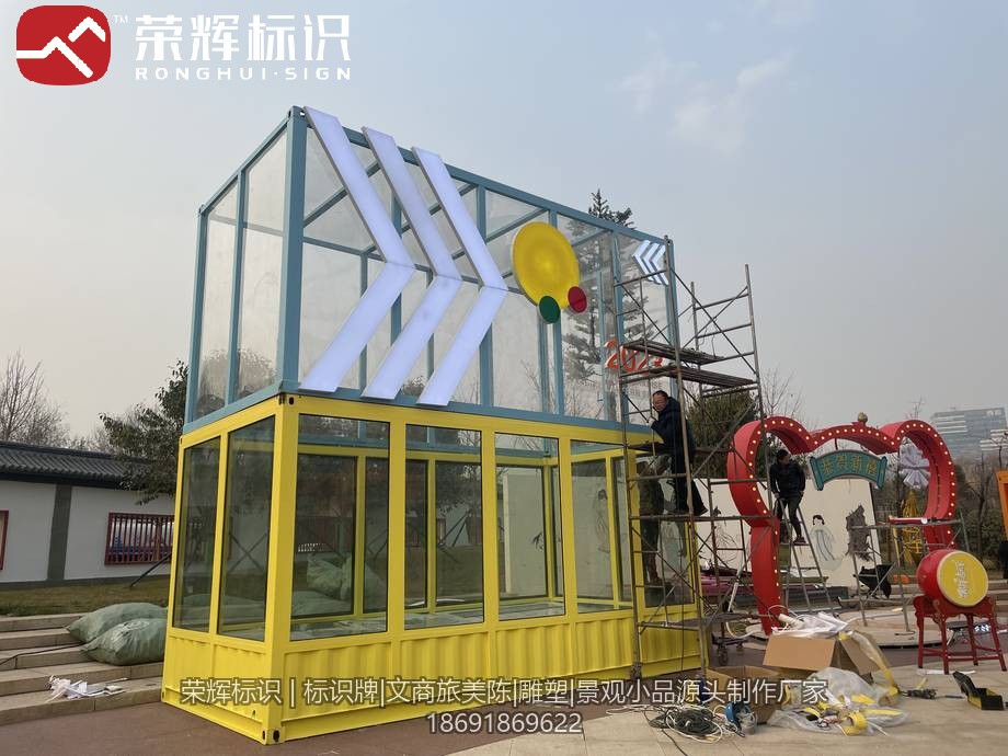 白天现场组装的集装箱美陈装置，黄色箱体叠加浅蓝框架与装饰造型件