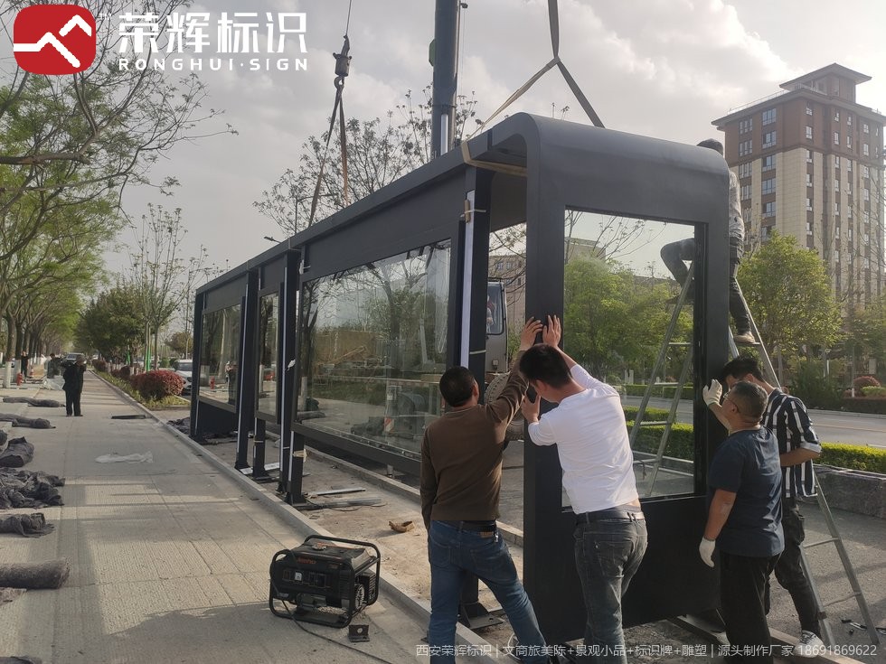 鄠邑公交候车亭吊装现场,吊带悬吊深灰金属框架带玻璃面板的候车亭,工人在旁配合