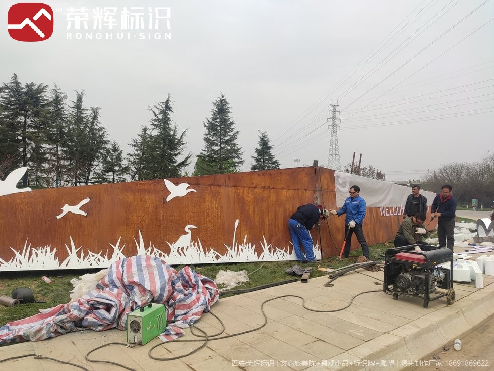 鄠邑耐候钢镂空景墙现场安装,锈色钢板配白色飞鸟与芦苇镂空图案,工人在旁配合定位