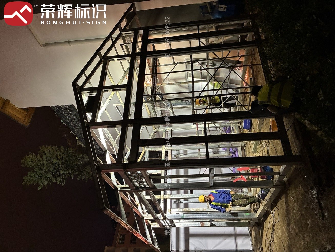 夜间施工时段内的美陈装置安装现场，装置面板从内部透出亮光，工人在脚手架上进行固定作业